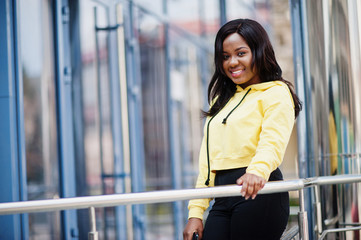 Hipster african american girl wearing yellow hoodie posing at street against office building with blue windows.
