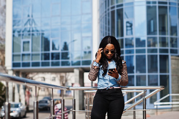 Hipster african american girl wearing sunglasses, jeans shirt with leopard sleeves, hold mobile phone, posing at street against modern office building.