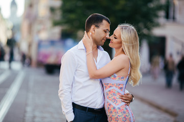 Lovely couple walking around the block. Dark-haired man in a white shirt hugging a blonde in a beautiful dress