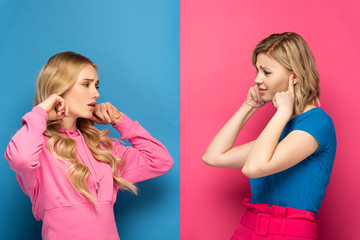 Skeptical blonde sisters covering ears while looking at each other on pink and blue background