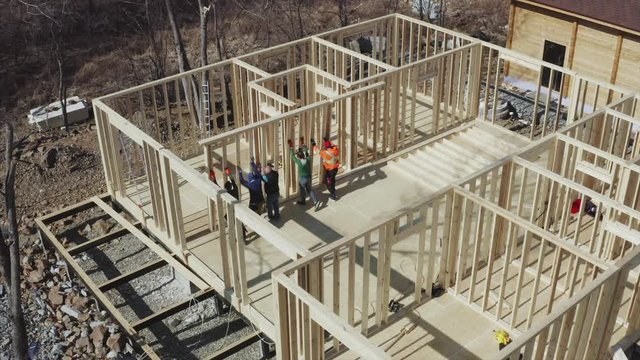 Aerial view of builders team lifting the wooden wall on the place. Residential frame house under construction. Clear sunny day