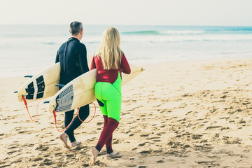 Rear view of couple with surfboards walking on beach. Back view of man and woman in wetsuits holding surfboards and walking on sea coast at summertime. Surfing concept