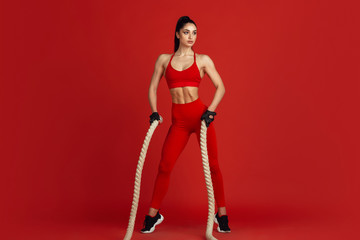 Posing strong. Beautiful young female athlete practicing in studio, monochrome red portrait. Sportive fit brunette model with ropes. Body building, healthy lifestyle, beauty and action concept.