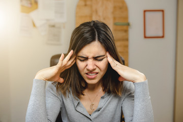 portrait of a young woman with headache sitting on a desk chair while working in office.