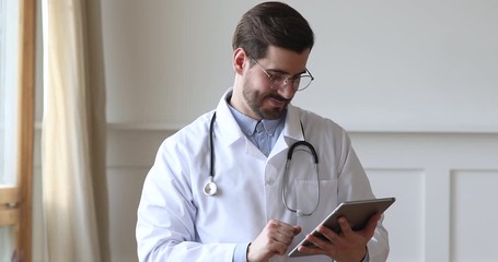 Head shot happy young male doctor in eyeglasses wearing white uniform with stethoscope, using digital computer tablet. Smiling professional general practitioner doc consulting clients patients online.