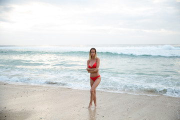 A beautiful sexy girl model relax on white sand beach, in red bikini swimsuit, with a tanned body, long blonde hair, magnificent breasts and puffy lips. Healthy nature, ocean waves, clouds on Bali