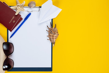 A reminder of what not to forget to take with you on a trip: tablet with pen, paper airplane, passports, sunglasses, hat, shell and starfish on a yellow background