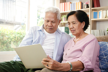 Fototapeta premium An elderly Asian couple sitting on a sofa watching media online on a laptop In the living room at home Both are happy in life after retirement. Concept of health insurance, Social distance