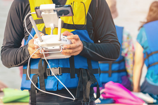 Young man in blue and yellow life vest controlling a drone to take pictures of ocean coast with tourists while traveling on a raft in the sea. Hands holding drone remote controller.