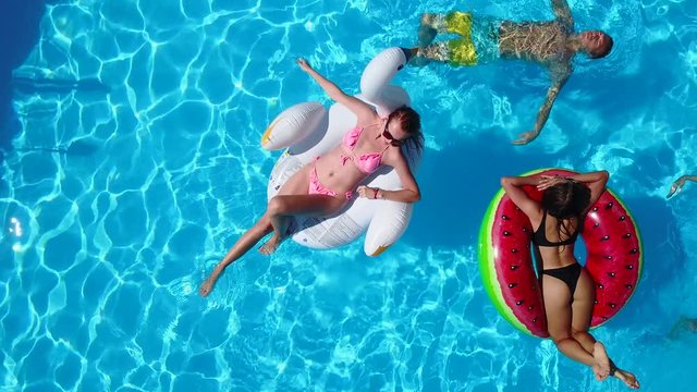 Aerial. Friends chilling in swimming pool with inflatable flamingo, swan, mattress. Happy young people bathe on floating mattresses in luxury resort. View from above. Girls in bikini sunbathing in sun