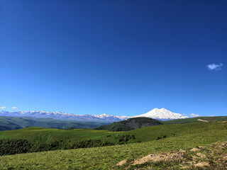 Great nature mountain range. Amazing perspective of caucasian snow mountain or volcano Elbrus with green fields, blue sky background. Elbrus landscape view - the highest peak of Russia and Europe