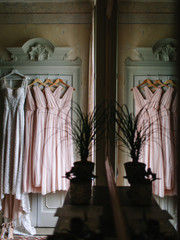 A white wedding dress and bridesmaids dresses in pink are hanging on an old vintage wardrobe of light green color. On the mosaic floor are the shoes of the bride. And an open window