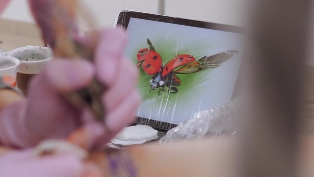 Close-up of a tablet with a picture of a ladybug on a table in a tattoo parlor, in the foreground a tattoo artist puts a tattoo on the skin of a young girl.