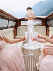 Girls in pink dresses clink glasses with white glasses with prosecco against the background of a bride standing in a white wedding dress. Sitting in a boat ride on a lake in Italy.