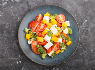 Vegetarian salad with broccoli, tomatoes, feta cheese, and pumpkin on a blue ceramic plate on a black concrete background, top view