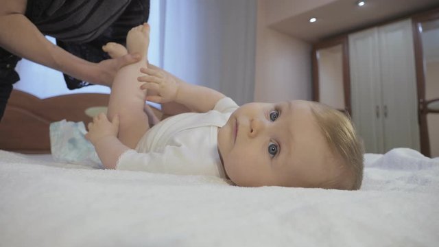 Careful mother changes baby diaper at home. Cute toddler with blue eyes lies on a diaper in bedroom. Mom and toddler concept of a happy childhood. Low-angle shot in close-up