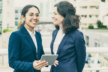 Obraz premium Happy businesswomen with digital tablet. Cheerful professional female colleagues standing with digital tablet on urban city street. Wireless technology concept