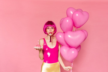 Image of woman blowing in party horn while holding balloons and cake