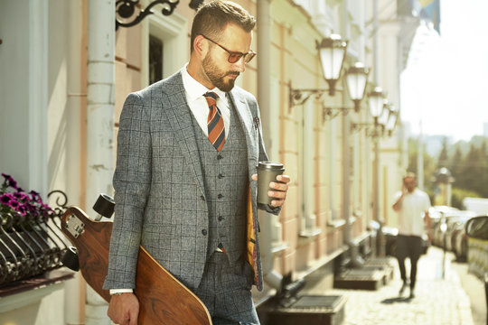 Fashionable European business man in perfect suite walk on the street, using skateboard longboard and suit man, coffee take away and smile me