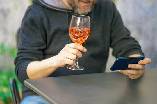 Man Holding A Spritz Alcoholic Cocktail In His Hands Making An Online Celebrative Toast With His Friends On A Happy Hour Video Call Safe In The Courtyard. Quarantine Coronavirus Aperitif Party.