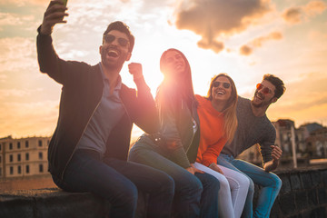 Fototapeta premium Group of happy young influencers people taking a selfie sitting on a bench outdoors in the sunset. Real backlight flare. Leisure activities lifestyle concept.