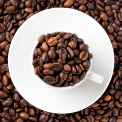 Small white cup with coffee beans and saucer on a layer of full roasted coffee beans in a full frame background texture. Top view.