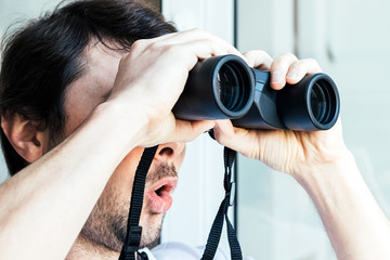 Handsome man with a surprised face looking through binoculars at the window with a view of the...