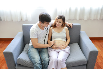 Obraz premium Top view handsome caucasian husband using medical stethoscope to listening his asian pregnant belly wife while sitting on sofa in living room with love