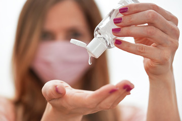 Young woman with home made pink cotton face mouth virus mask dropping anti bacterial alcohol rubbing gel on hand, detail to white bottle, blurred face in background. Coronavirus covid 19 prevention