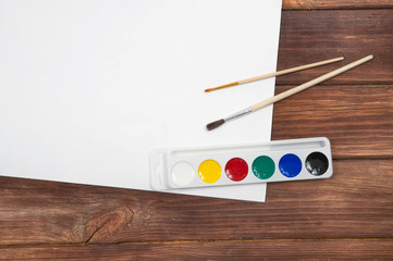 multi colored children's paints with two brushes in the right part of the image on a white blank sheet of paper on a wooden table top view