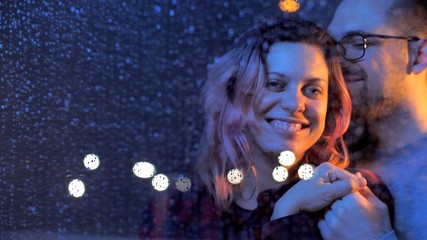 Husband hugs his smiling wife on the shoulders. Portrait of people looking at the camera. Married Loving couple man and woman hugs and look through a window wet from rain. At night stay home