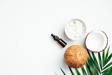 Coconut essential oil bottle with sliced coconut in bowl, tropical palm leaf, half of coconut on white background. Flat lay, top view. SPA natural organic cosmetic for skin care, body treatment