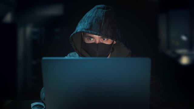 Hacker hooded and hide face with mask typing code on keyboard late night in dark room