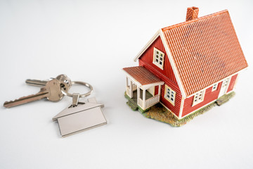 Red miniature house standing on white office desk next to home keys. House, real estate, buyer and sold concept.