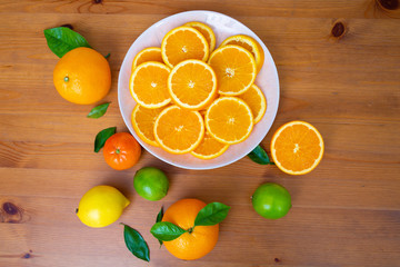 Big white ceramic plate with sliced oranges. Fresh mandarins, limes, lemon and citrus with leaves on wooden desk near bowl. Top view. Studio shot. Nutrition and vegetarian concept
