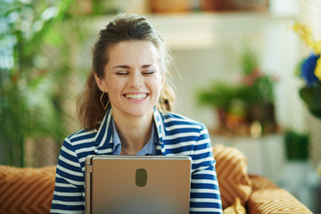 smiling trendy housewife hugging laptop while sitting on sofa