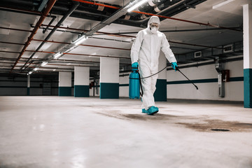 Worker in sterile uniform and mask walking trough underground garage and sterilizing surface. Protection from corona virus / covid-19 concept.