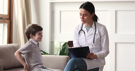 Friendly young female pediatrician consulting small cute kid boy patient at home. Little child sitting on couch, answering questions of pleasant family doctor general practitioner at checkup meeting.