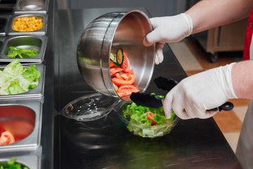 Cook preparing a healthy, vegetarian, to-go salad in a fast-food restaurant. Process of putting sliced vegetables in a plate from a bowl.