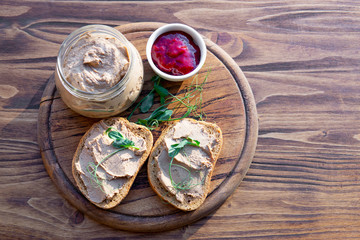 Homemade chicken liver pate (goose, rabbit) on bread and in a jar, cranberry sauce, wooden chopping board, rustic table, natural light. Copy space, selective focus.