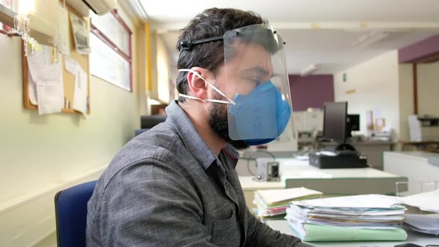 Office worker with plastic protective screen and face mask working on computer