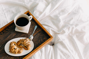 Breakfast in bed: top view of a wooden tray on white bed sheet, with two croissants and cup of coffee.
