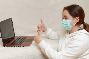 A young girl in a mask lies at home on a sofa with a computer and communicates via skype with...