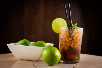 alcoholic mojito with mint and a bowl full of limes on dark background. Mojito with lime and mint with straw in a cup of glass. General shot.