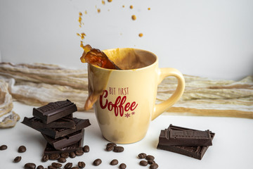 View of a coffee splash in a light beige coffee cup with lettering surrounded by chocolate bars and coffee beans