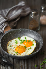 Fried eggs with green chives chopped for breakfast. Two fried eggs in a small pan on the dark wooden background. Rustic