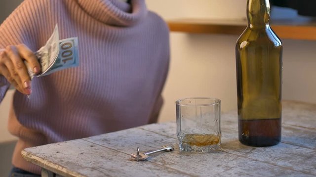 Sad woman in jeans and pink sweater comeback home, puts dollars and keys on the table and pours whisky on her glass from the green bottle because of the problems, female alcohol addiction