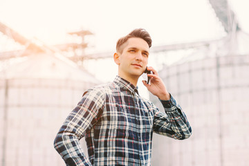 Portrait of young business man talking mobile phone while working at silage storage factory. Happy agriculture business manager holding cell phone in hand while standing against silos building © Artem