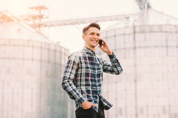 Portrait of happy agriculture industry business owner talking cell phone and smiling while standing against silos warehouse building. Young confident business man farmer holding mobile phone in hand