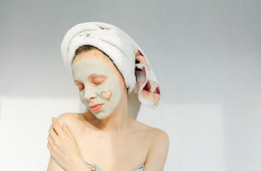 young woman with towel wrapped around her head applying face clay mask at the white background